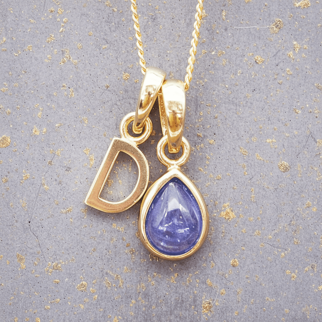 Gold necklace with 'D' initial pendant and teardrop-shaped blue december birthstone tanzanite gemstone on a grey background.