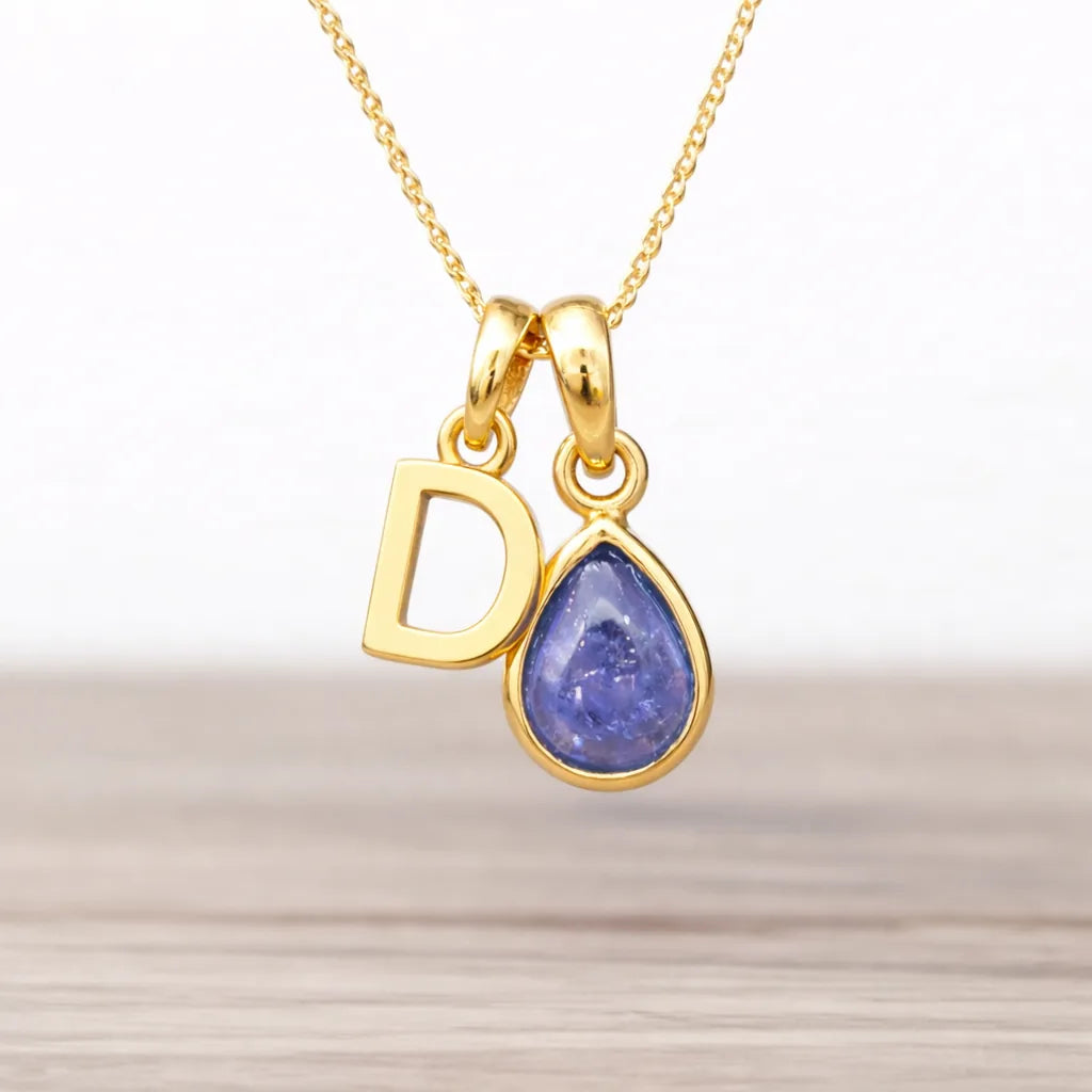 Gold necklace with a 'D' pendant and a teardrop-shaped purple tanzanite gemstone on a light wooden surface.