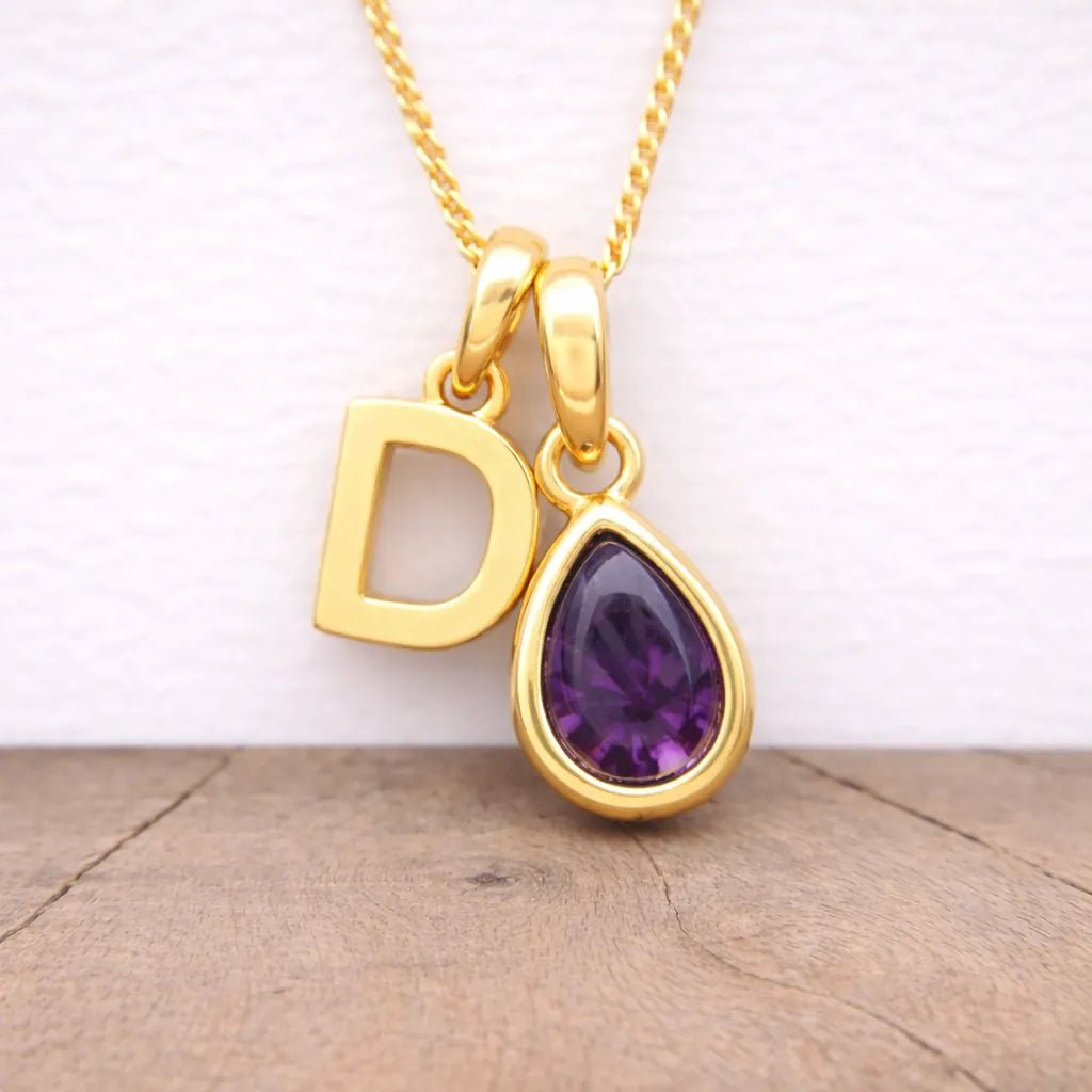 Gold necklace with 'D' pendant and purple teardrop gemstone on a wooden surface.