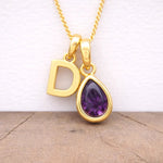 Gold necklace with 'D' pendant and purple teardrop gemstone on a wooden surface.