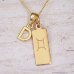 Gold initial D and Gemini zodiac bar necklace, close-up of the letter charm and engraved pendant on whitewashed wood