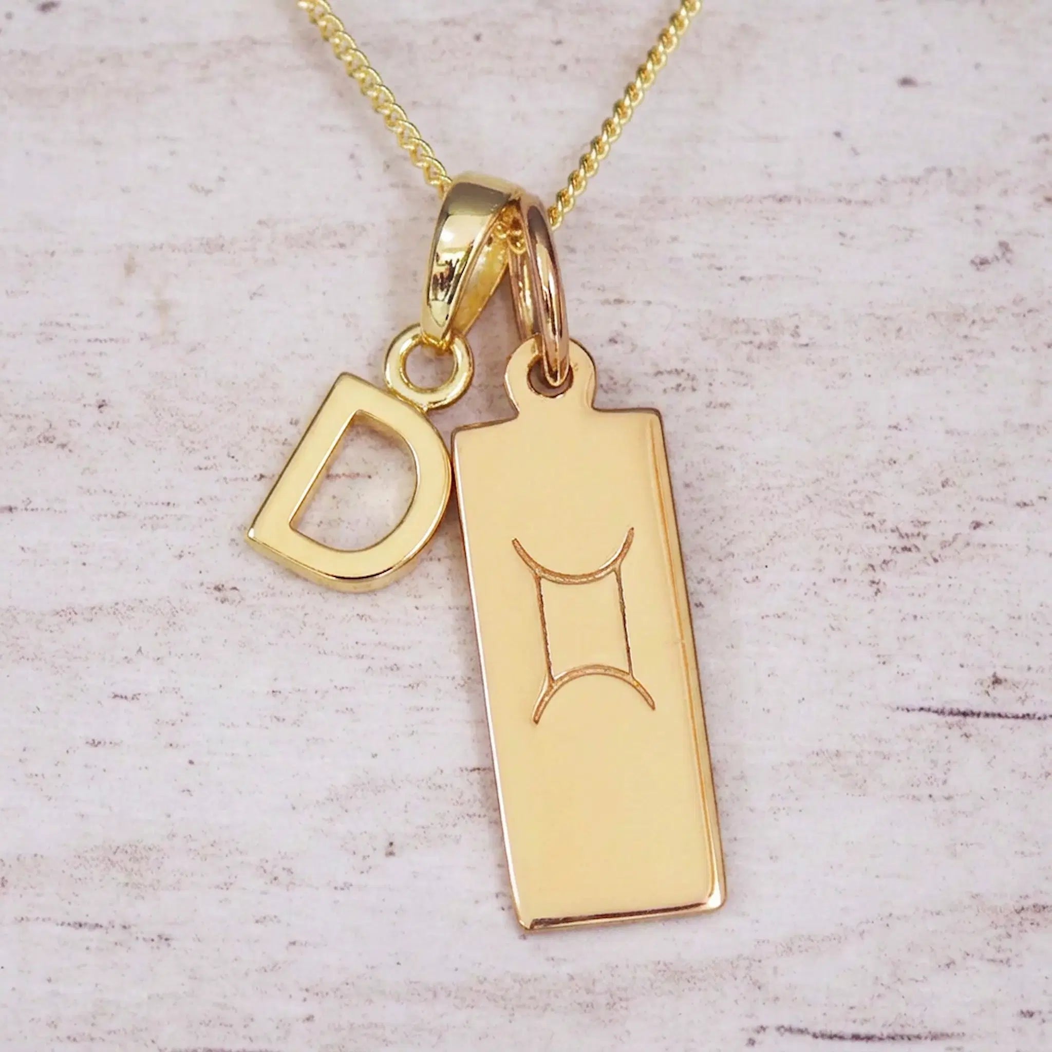 Gold initial D and Gemini zodiac bar necklace, close-up of the letter charm and engraved pendant on whitewashed wood