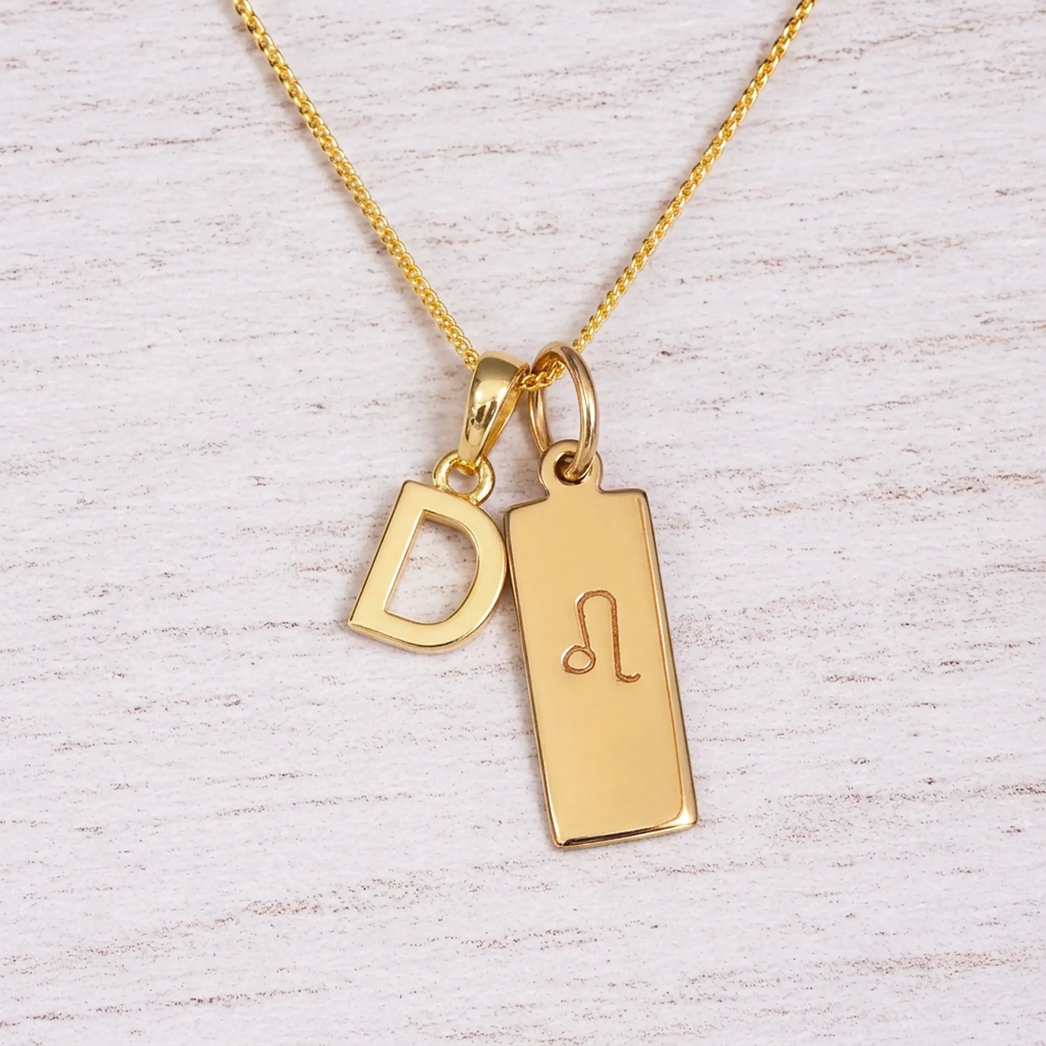 Gold initial and Leo zodiac necklace with a letter D pendant and engraved Leo bar on white wood, close-up
