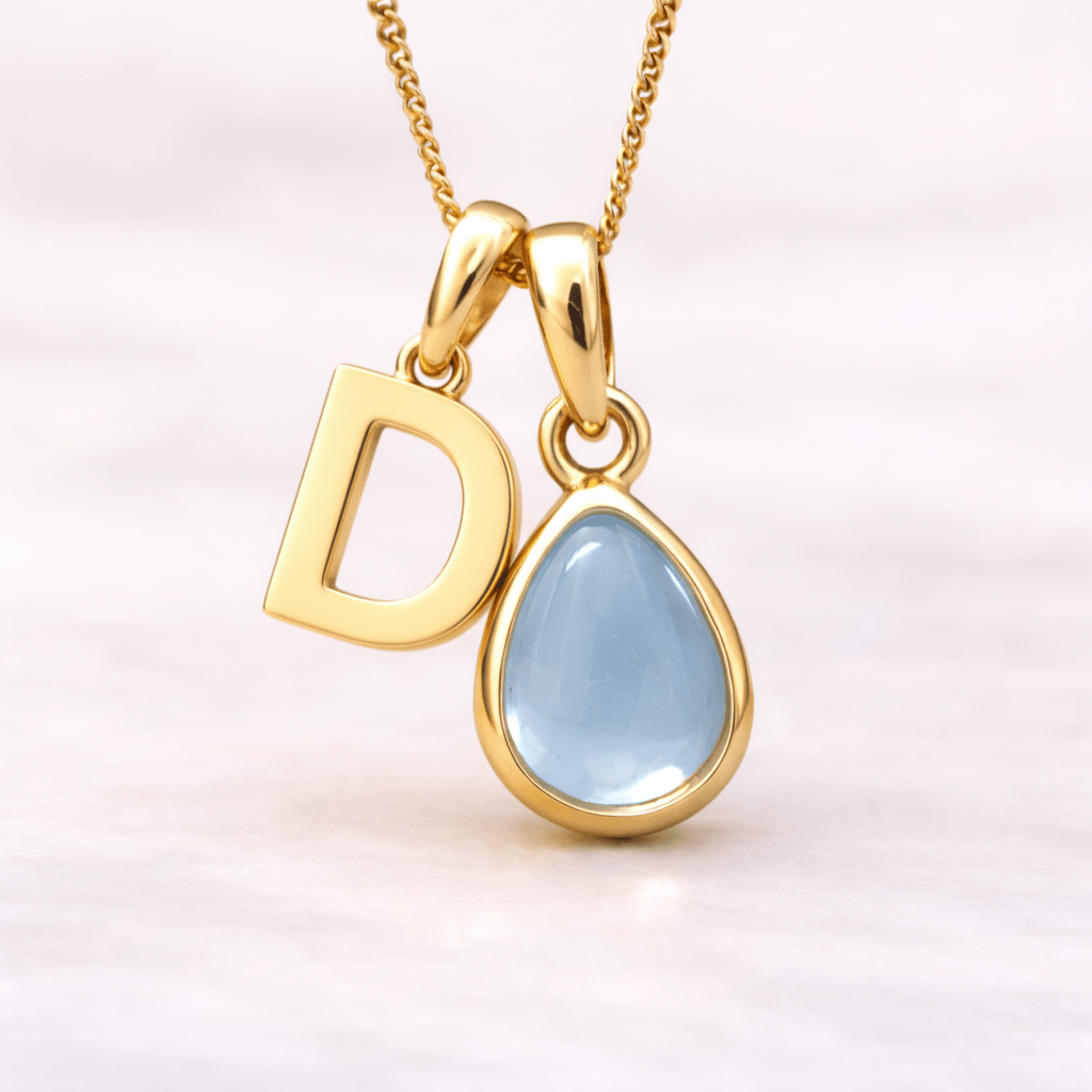 Gold necklace with 'D' pendant and teardrop-shaped blue topaz gemstone on a light pink background