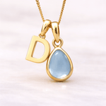 Gold necklace with 'D' pendant and teardrop-shaped blue topaz gemstone on a light pink background