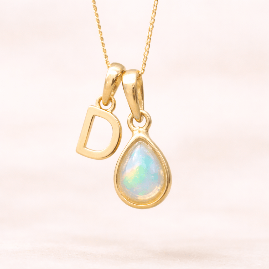 Gold necklace with a teardrop-shaped opal pendant and letter 'D' charm on a light pink background