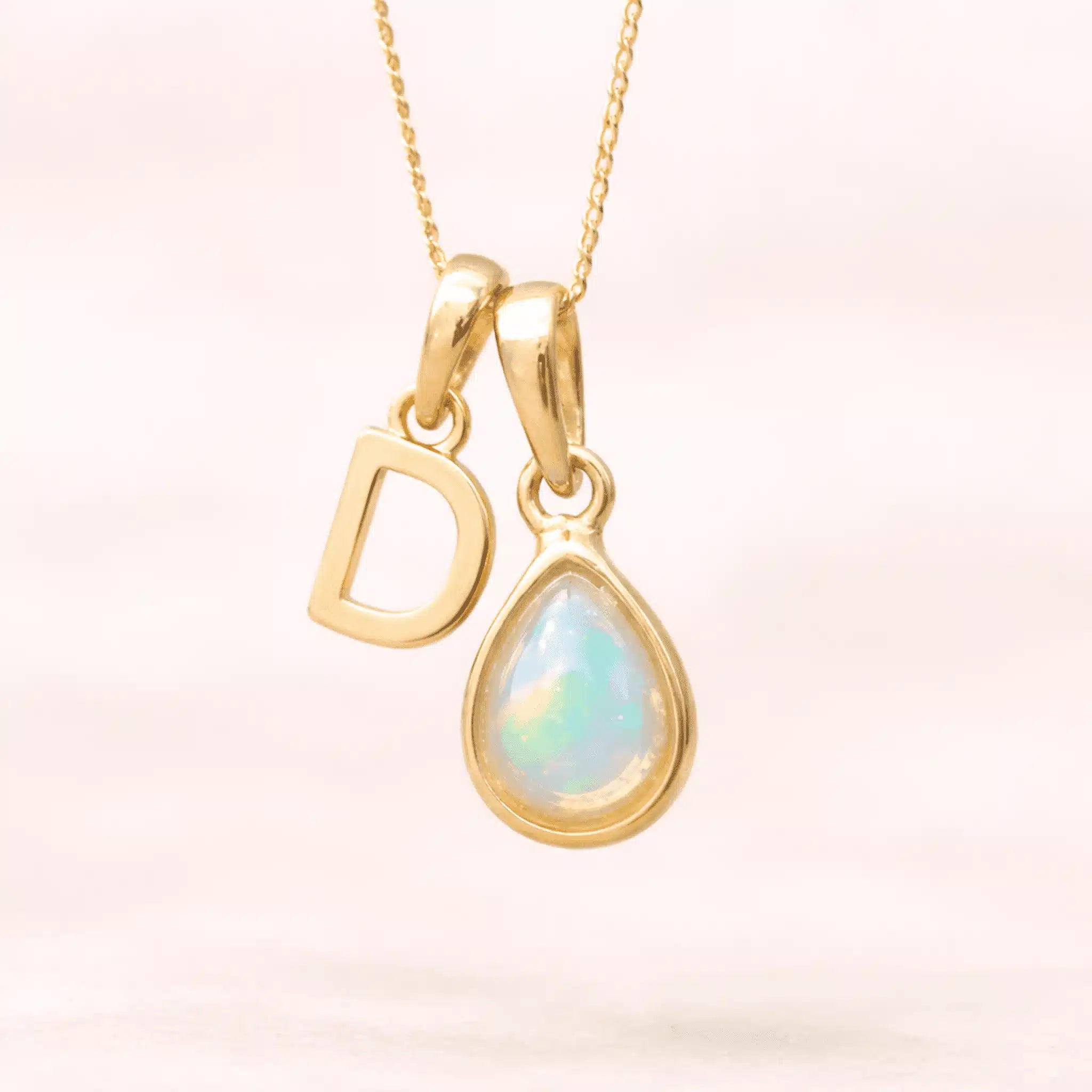 Gold October birthstone necklace with a pear-shaped natural opal pendant and letter 'D' charm with a light background.