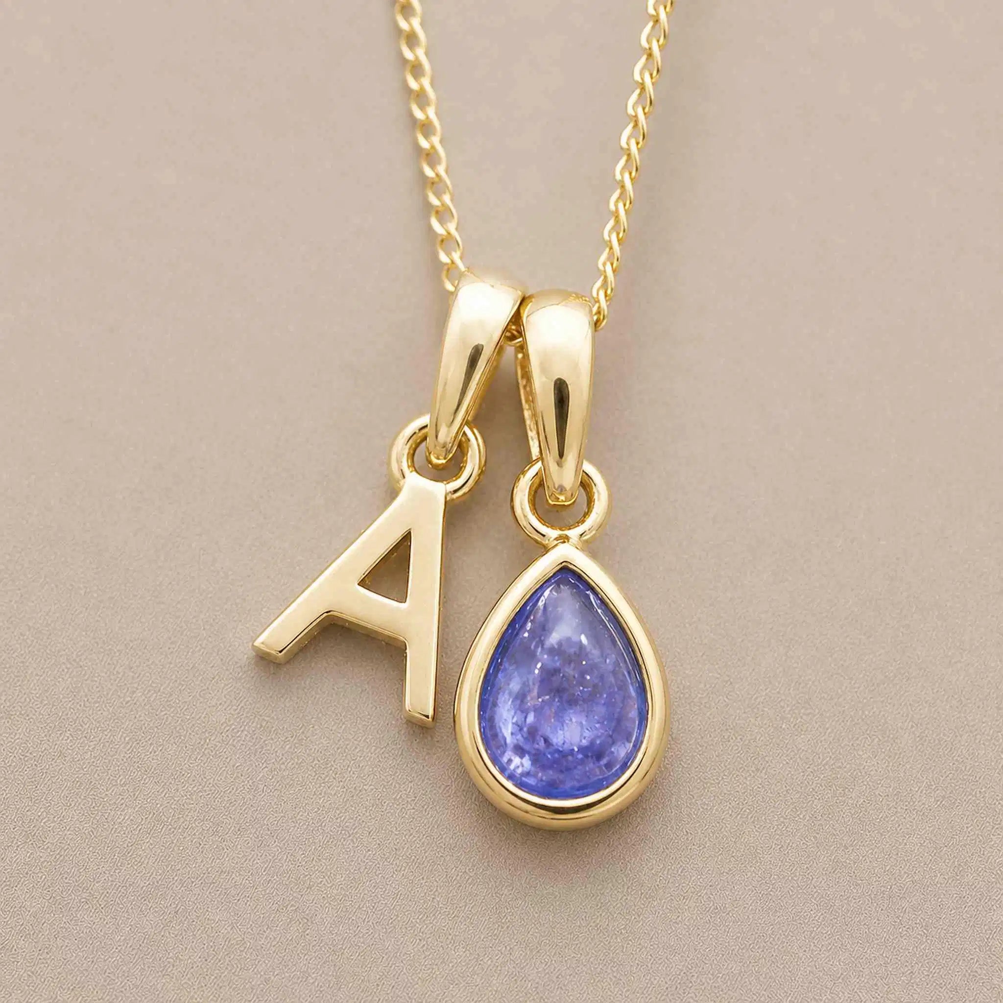 Gold December birthstone necklace with a pear-shaped natural tanzanite pendant and letter 'A' charm with a grey background.