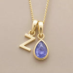 Gold fine chain necklace with a z-shaped pendant and a december birthstone purple teardrop tanzanite pendant on a beige background