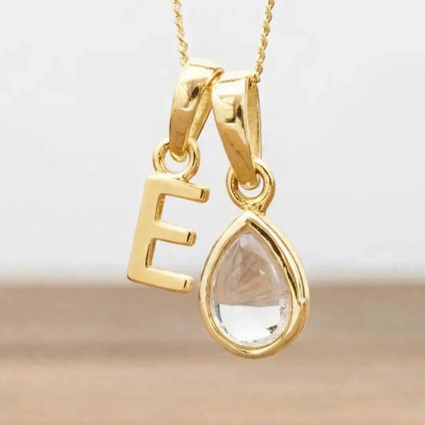 Gold necklace with 'E' pendant and teardrop-shaped herkimer quartz crystal on a light background