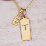 Gold initial E and Aries zodiac bar necklace, close-up of the letter charm and engraved pendant on whitewashed wood