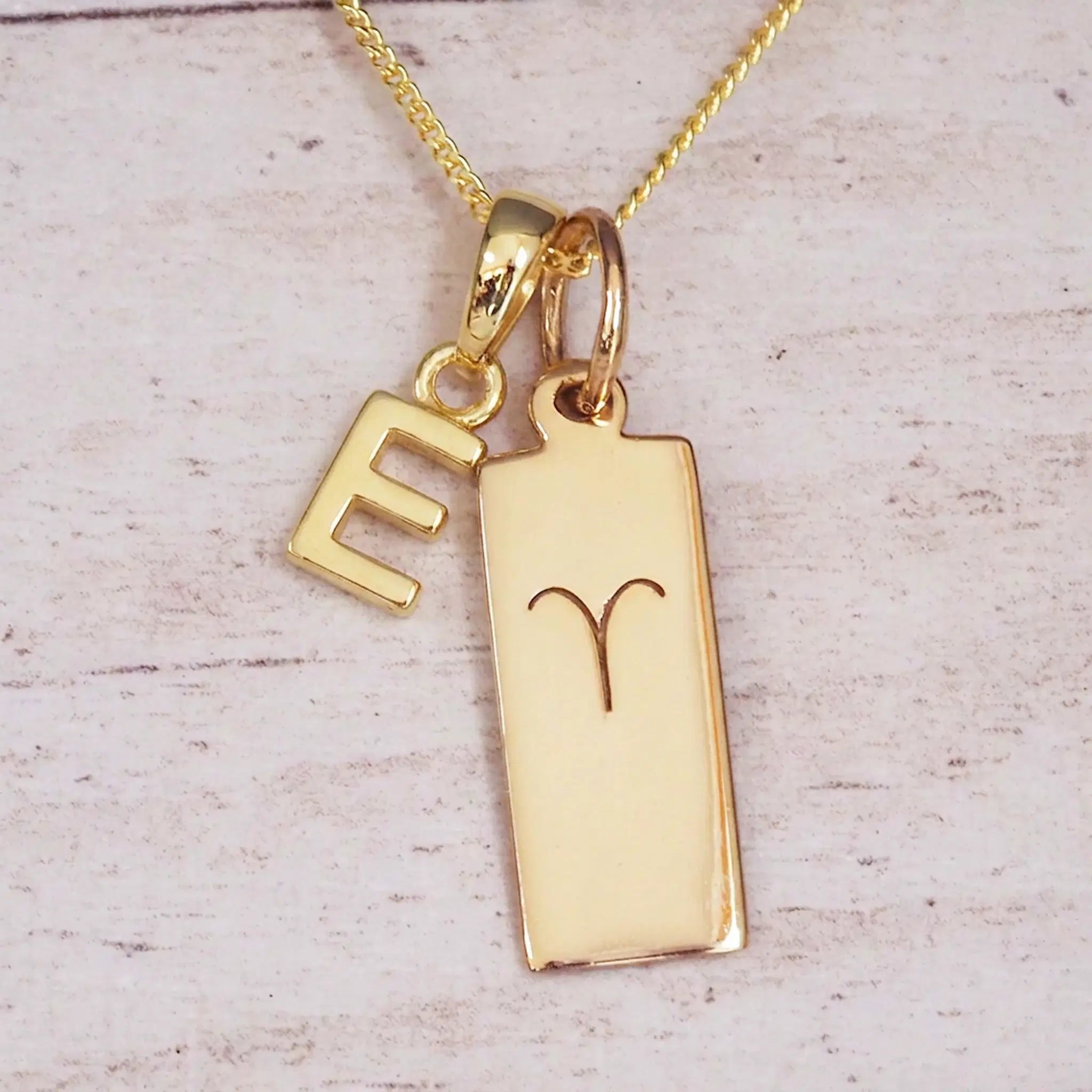 Gold initial E and Aries zodiac bar necklace, close-up of the letter charm and engraved pendant on whitewashed wood