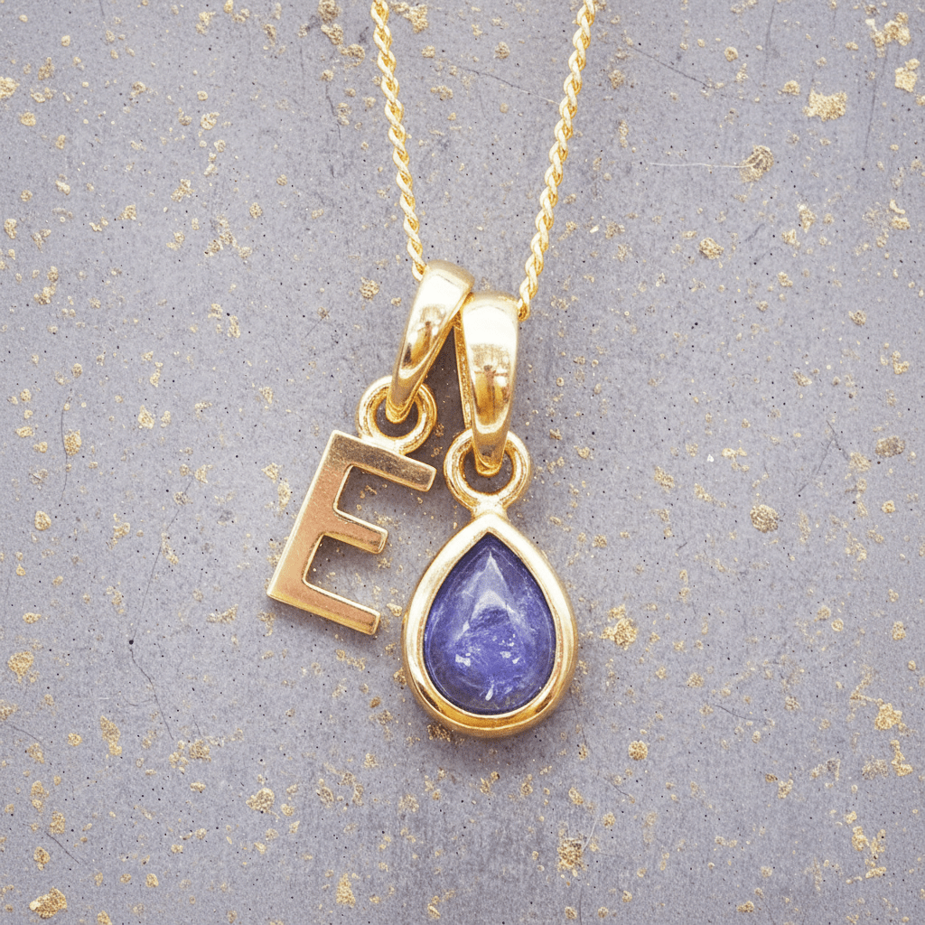 Gold necklace with 'E' pendant and purple teardrop tanzanite pendant on gray background