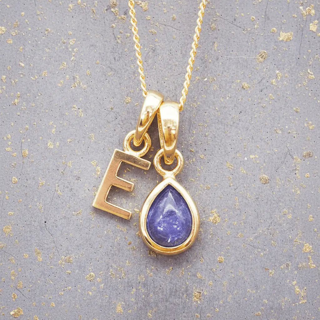 Gold December birthstone necklace with 'E' initial pendant and a natural tanzanite gemstone pendant on a grey background.