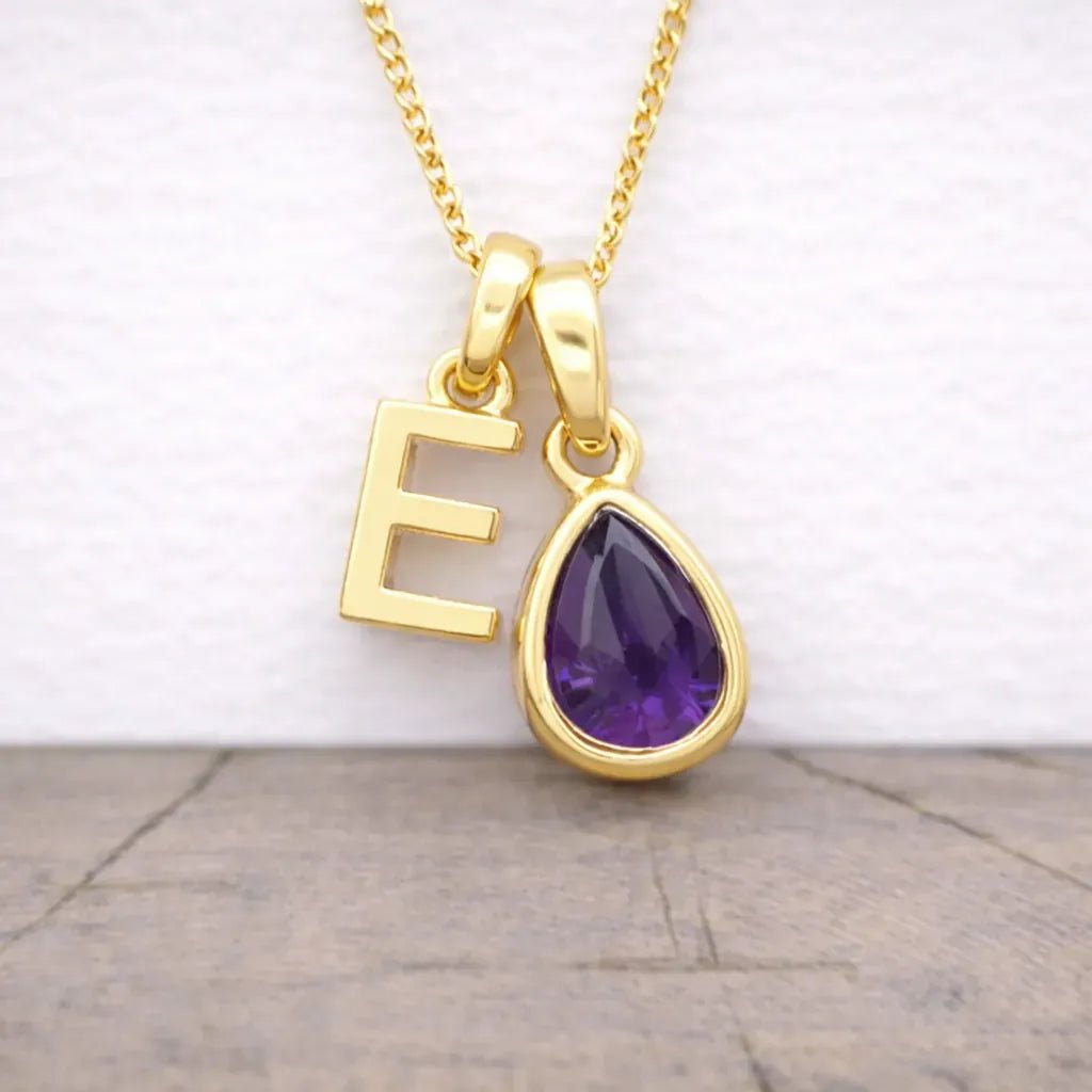 Gold necklace with 'E' pendant and purple teardrop gem on a wooden surface