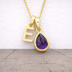 Gold necklace with 'E' pendant and purple teardrop gem on a wooden surface