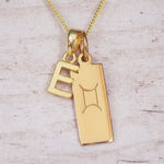 Gold initial E and Gemini zodiac bar necklace, close-up of the letter charm and engraved pendant on whitewashed wood