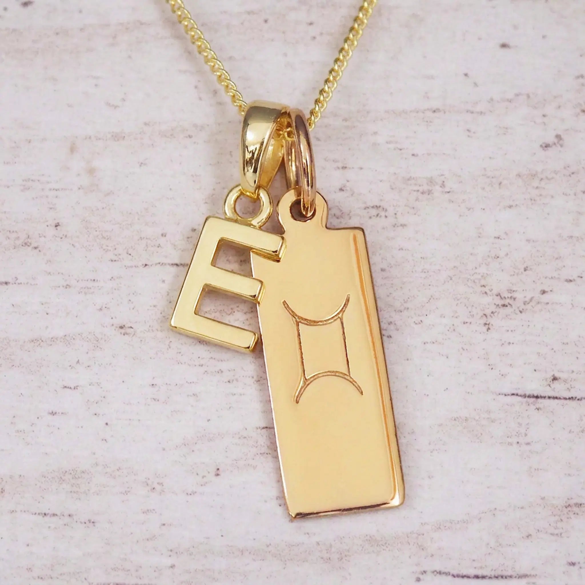 Gold initial E and Gemini zodiac bar necklace, close-up of the letter charm and engraved pendant on whitewashed wood