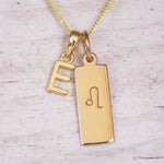 Gold initial and Leo zodiac necklace with a letter E pendant and engraved Leo bar on white wood, close-up