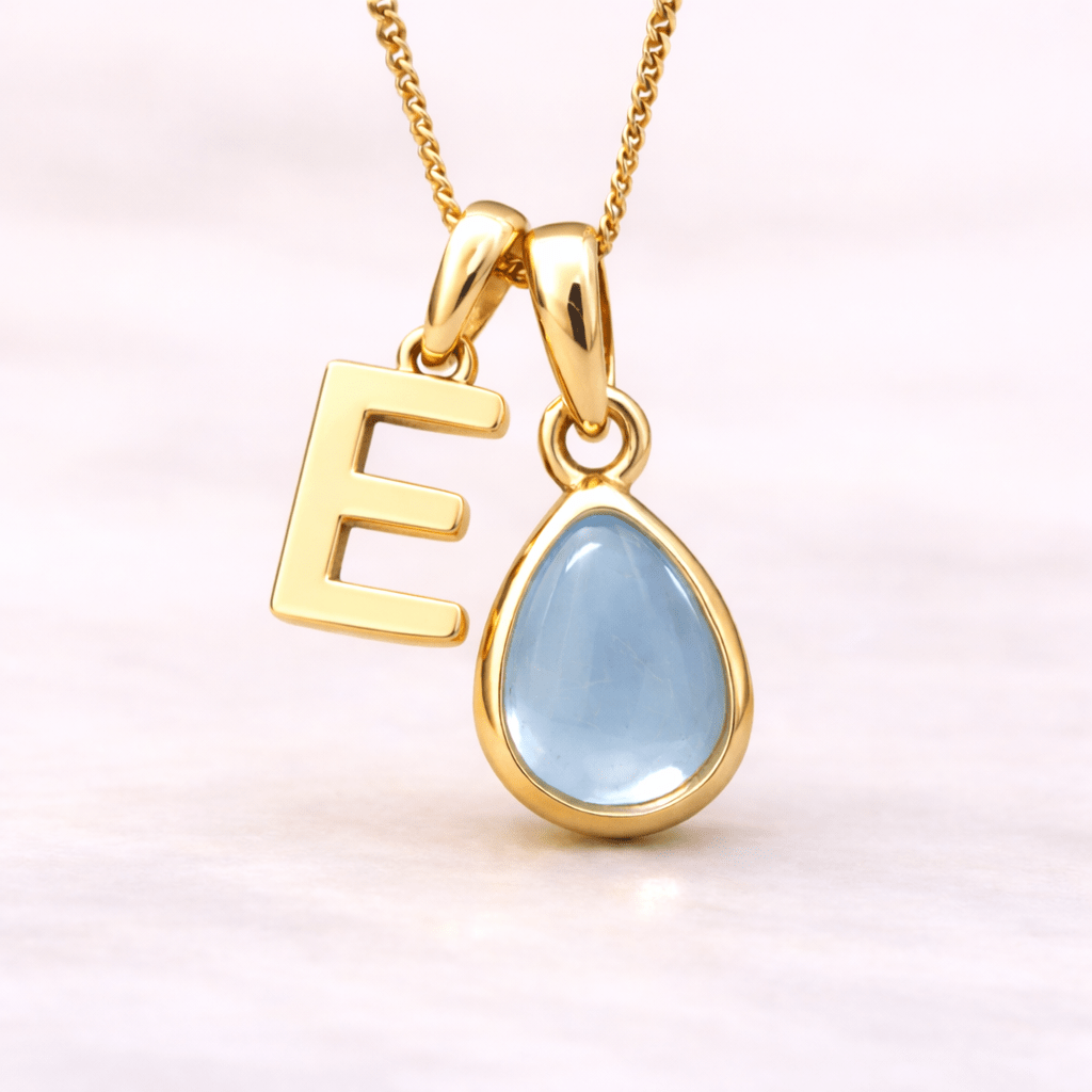 Gold necklace with 'E' pendant and teardrop-shaped blue topaz gemstone on a light pink background