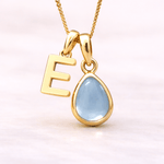 Gold necklace with 'E' pendant and teardrop-shaped blue topaz gemstone on a light pink background