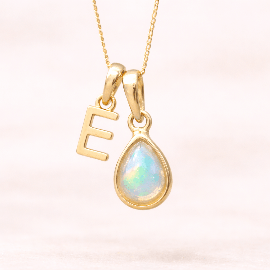 Gold necklace with a teardrop-shaped opal pendant and letter 'E' charm on a light pink background