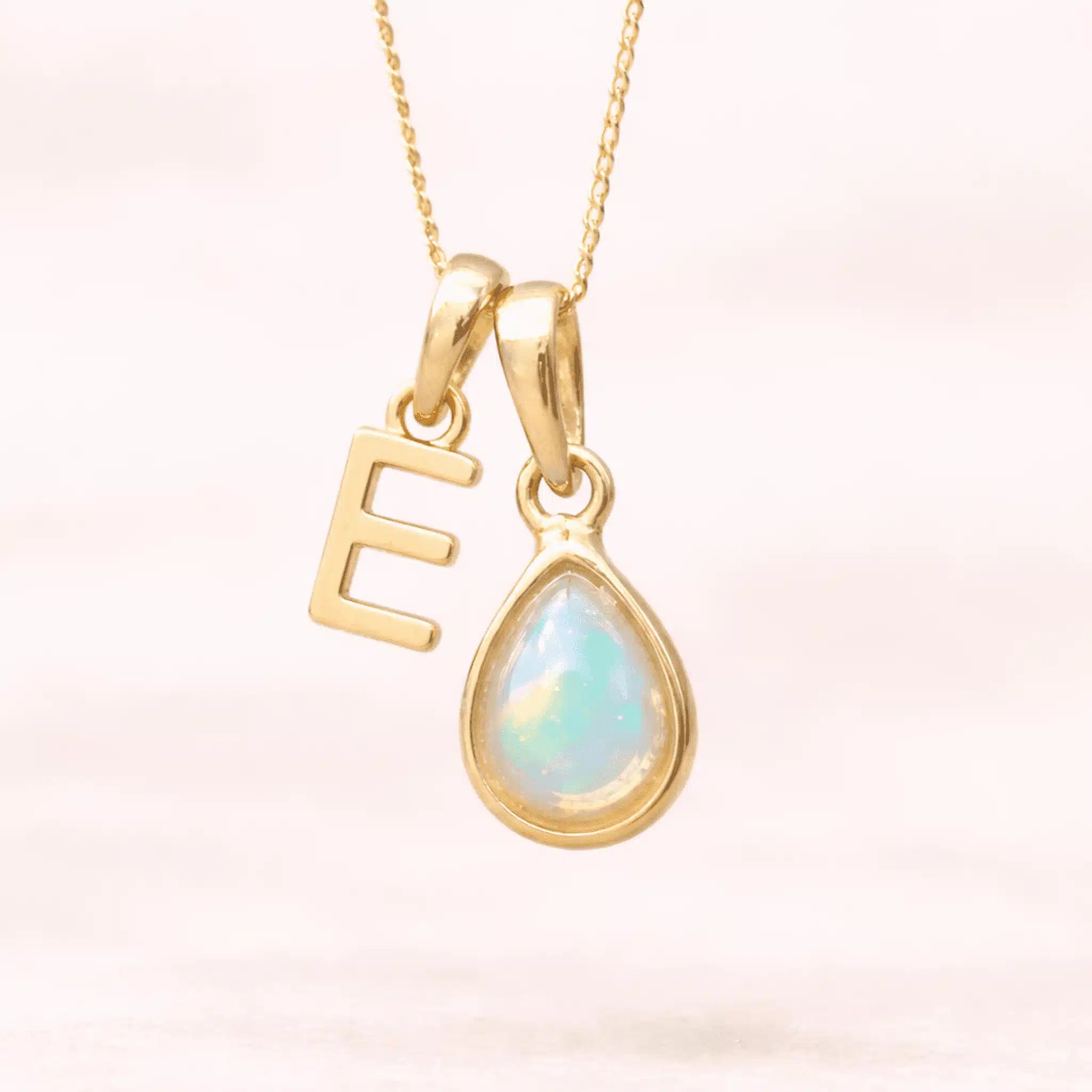 Gold October birthstone necklace with a pear-shaped natural opal pendant and letter 'E' charm with a light background.