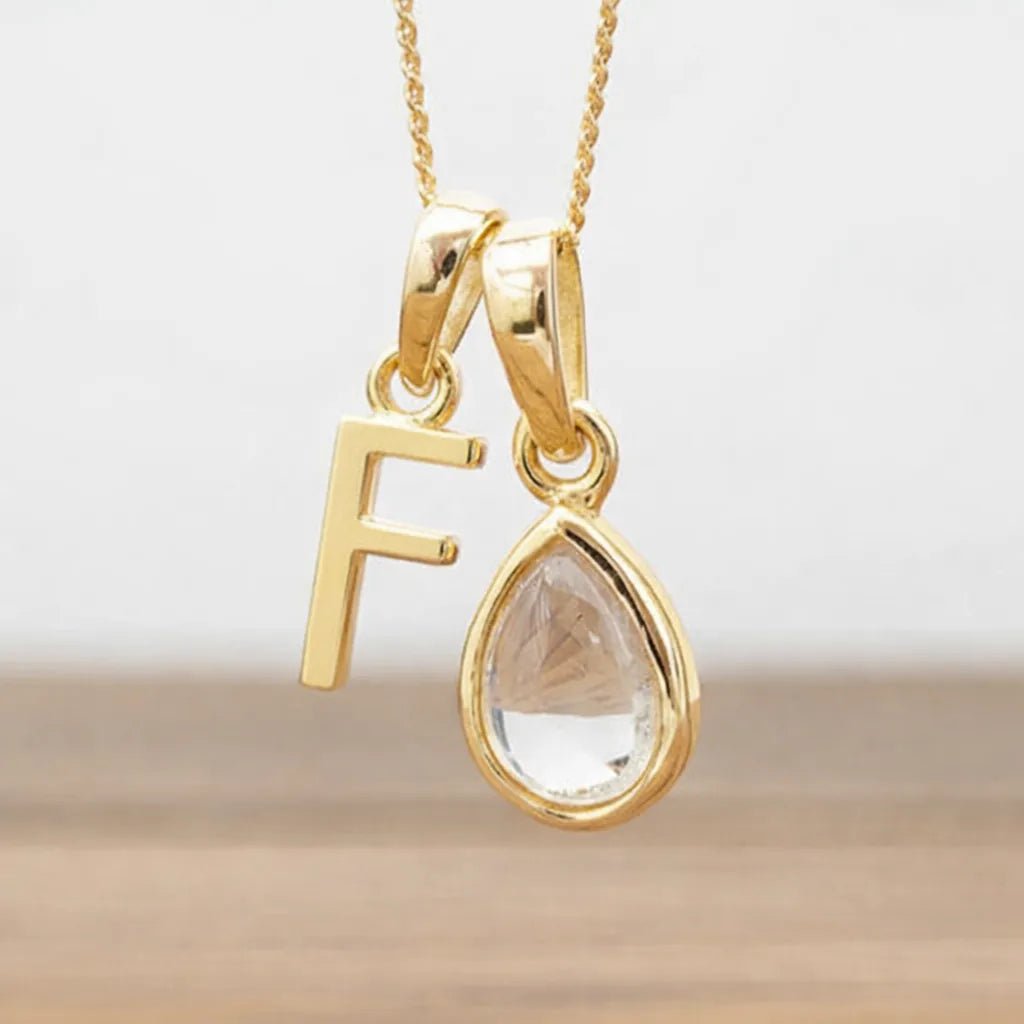 Gold necklace with 'F' pendant and teardrop-shaped clear herkimer quartz stone on a light background