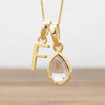 Gold necklace with 'F' pendant and teardrop-shaped clear herkimer quartz stone on a light background