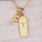 Gold initial F and Aries zodiac bar necklace, close-up of the letter charm and engraved pendant on whitewashed wood