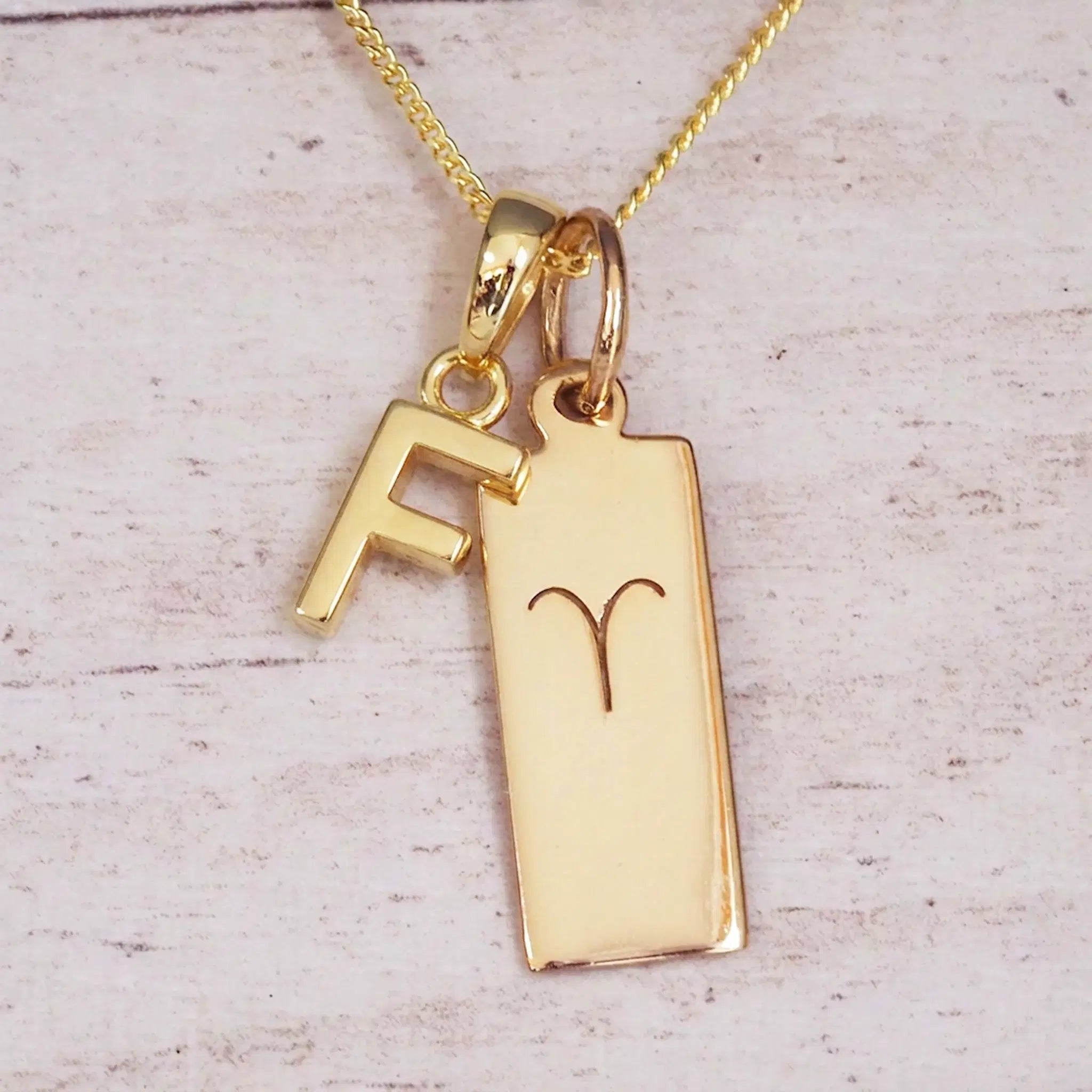 Gold initial F and Aries zodiac bar necklace, close-up of the letter charm and engraved pendant on whitewashed wood