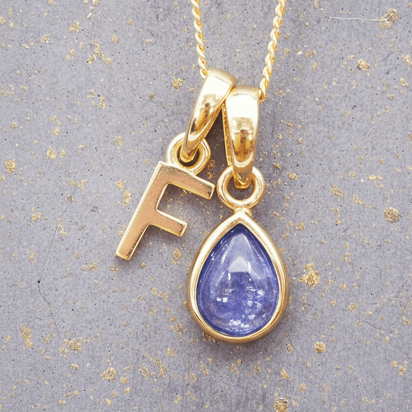 Gold necklace with a 'F' pendant and a teardrop-shaped blue tanzanite gemstone on a textured gray background.