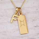 Gold initial F and Gemini zodiac bar necklace, close-up of the letter charm and engraved pendant on whitewashed wood