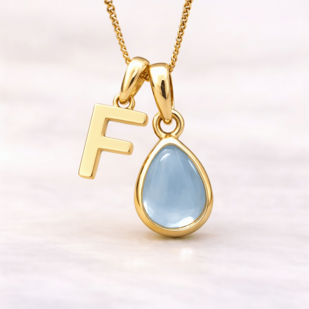 Gold necklace with a 'F' pendant and a teardrop-shaped light blue topaz gemstone on a white background