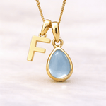Gold necklace with a 'F' pendant and a teardrop-shaped light blue topaz gemstone on a white background