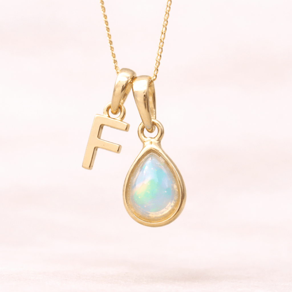 Gold necklace with a teardrop-shaped opal pendant and letter 'F' charm on a light pink background