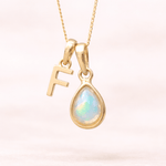 Gold necklace with a teardrop-shaped opal pendant and letter 'F' charm on a light pink background