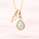 Gold October birthstone necklace with a pear-shaped natural opal pendant and letter 'F' charm with a light background.