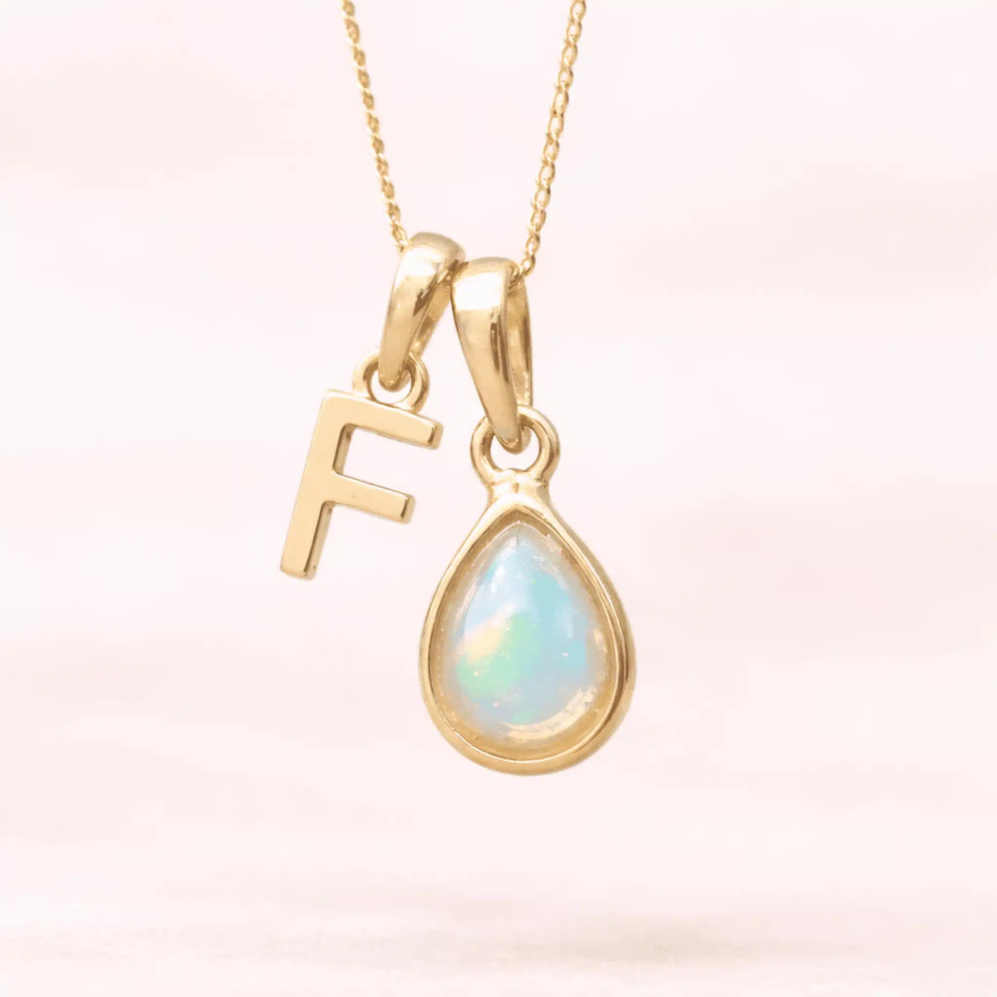 Gold October birthstone necklace with a pear-shaped natural opal pendant and letter 'F' charm with a light background.