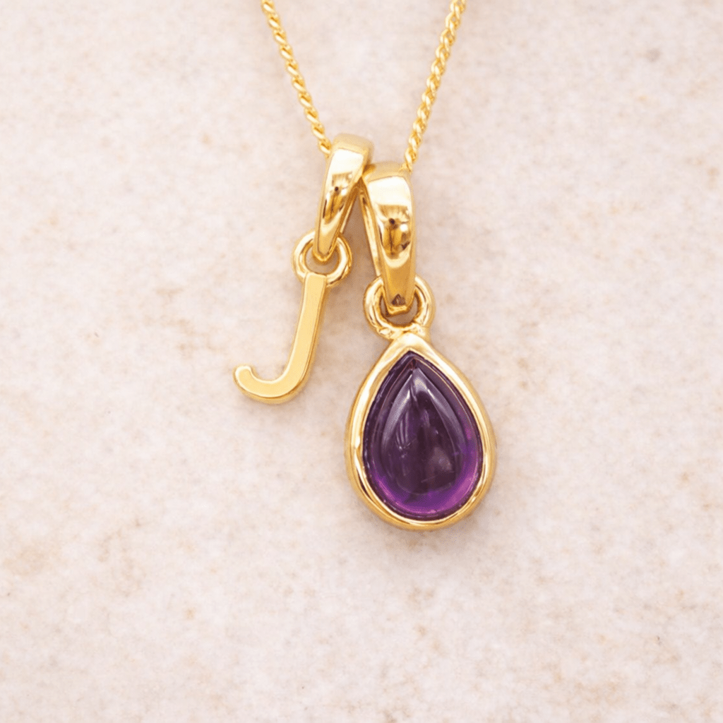 Gold initial J necklace with a purple teardrop amethyst pendant on a beige background