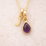 Gold initial J necklace with a purple teardrop amethyst pendant on a beige background