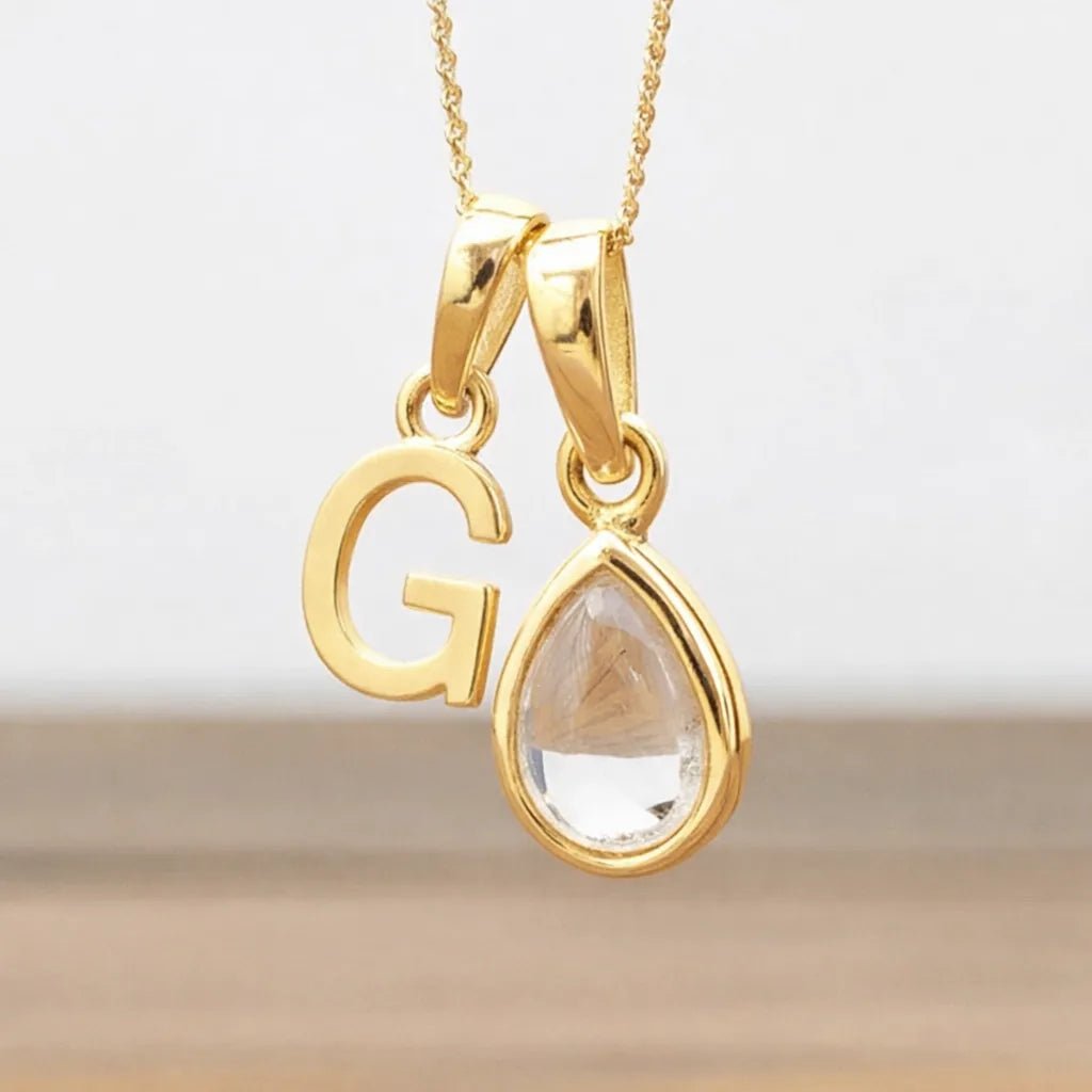 Gold necklace with 'G' pendant and clear teardrop-shaped gemstone on a light background