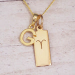 Gold initial G and Aries zodiac bar necklace, close-up of the letter charm and engraved pendant on whitewashed wood