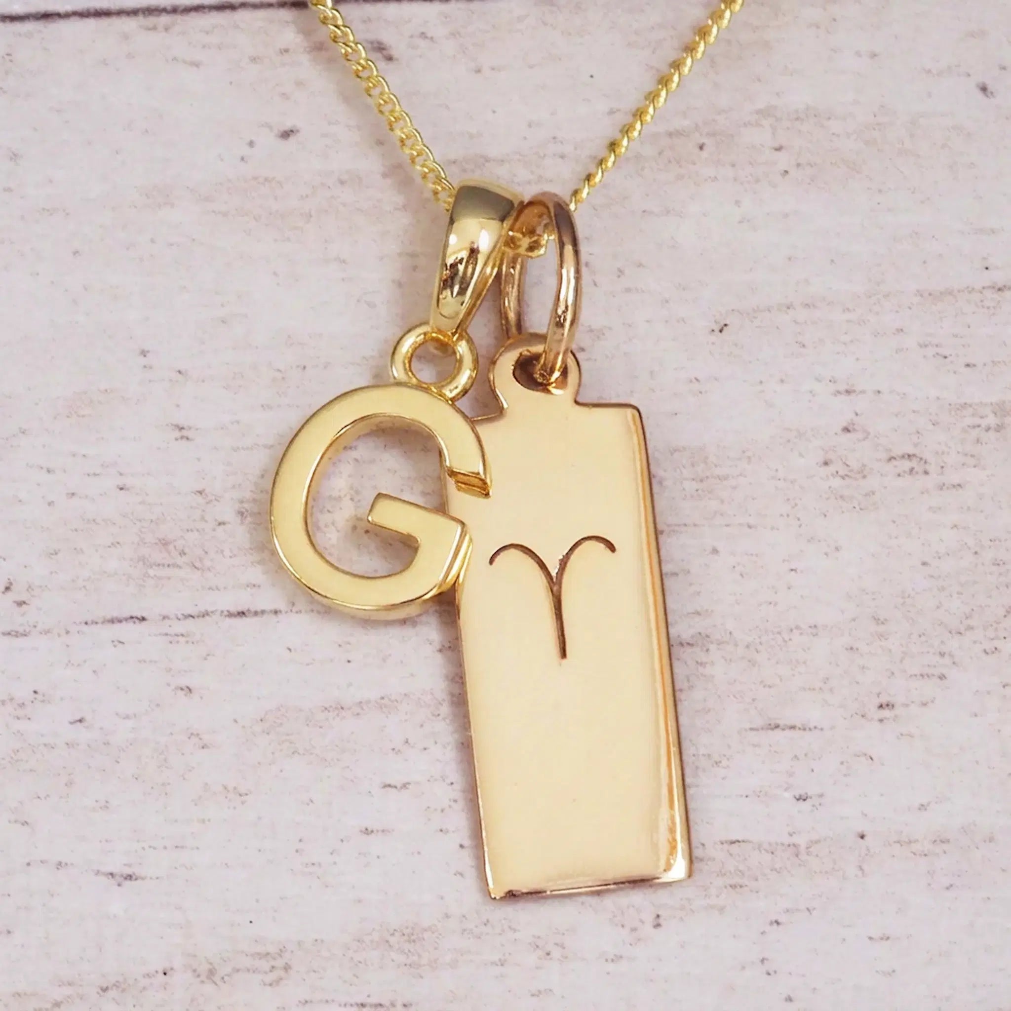 Gold initial G and Aries zodiac bar necklace, close-up of the letter charm and engraved pendant on whitewashed wood