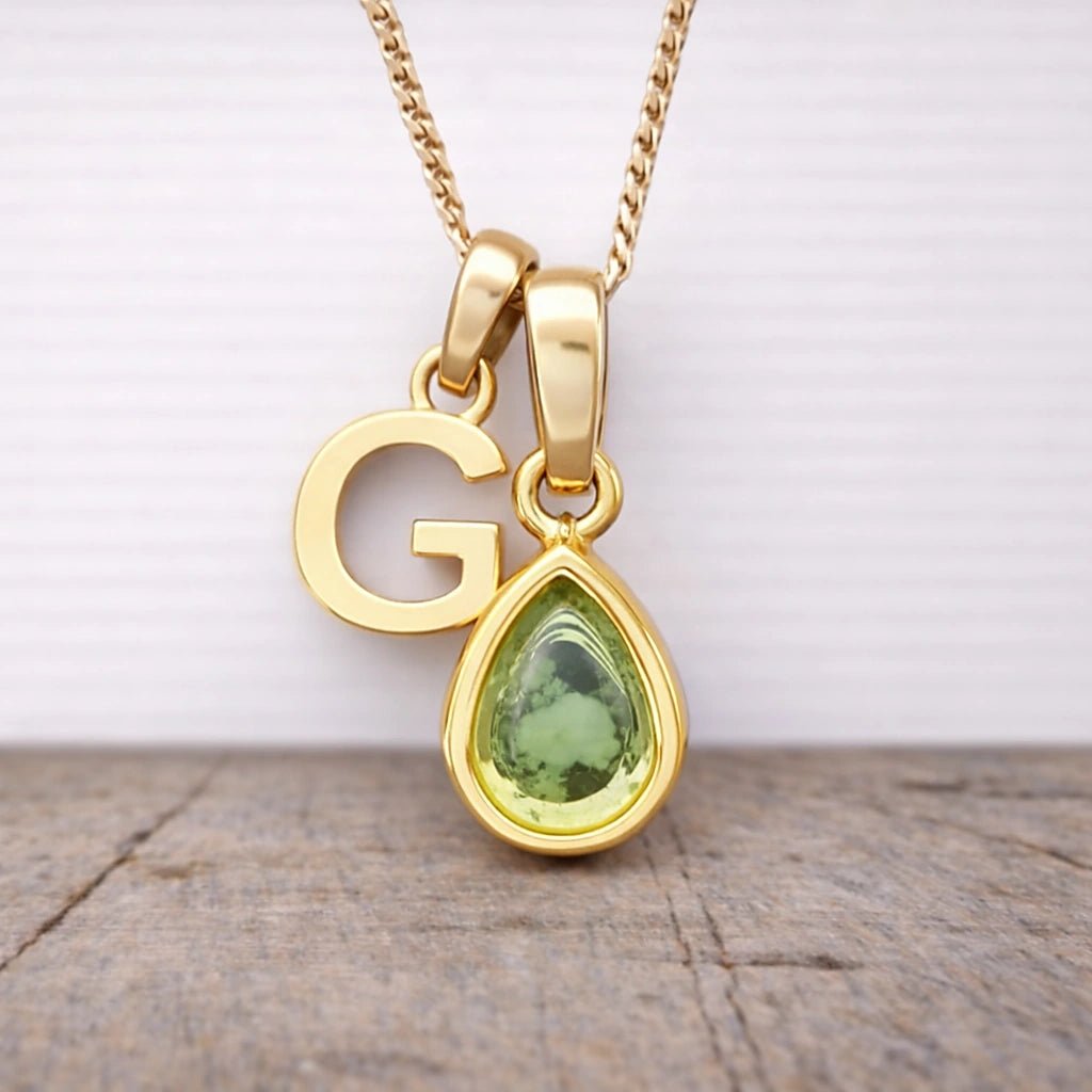 Gold necklace with a green teardrop pendant and letter 'G' on a wooden surface.