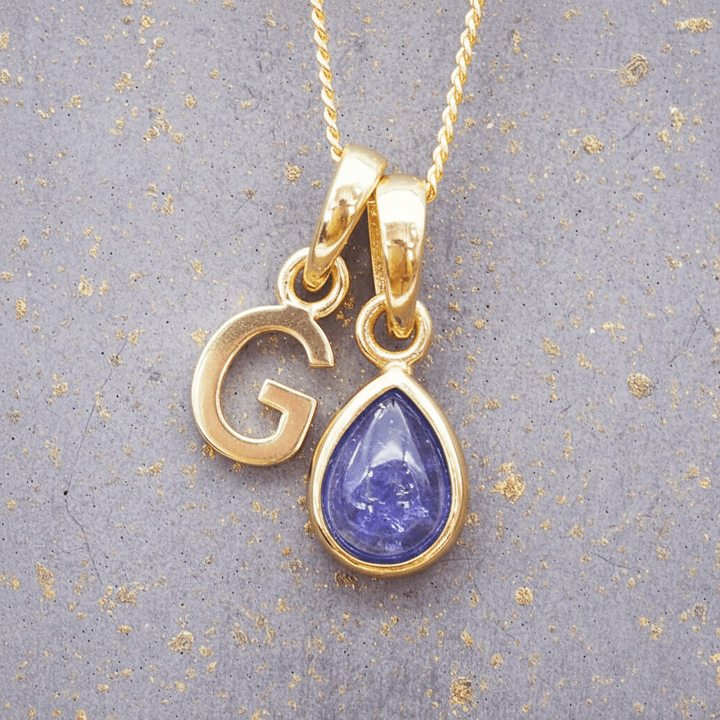 Gold necklace with 'G' pendant and teardrop-shaped purple tanzanite gemstone on a textured gray background.