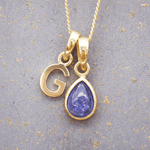 Gold necklace with 'G' pendant and teardrop-shaped purple tanzanite gemstone on a textured gray background.