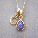 Gold December birthstone necklace with 'G' initial pendant and a natural tanzanite gemstone pendant on a grey background.