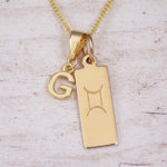 Gold initial G and Gemini zodiac bar necklace, close-up of the letter charm and engraved pendant on whitewashed wood