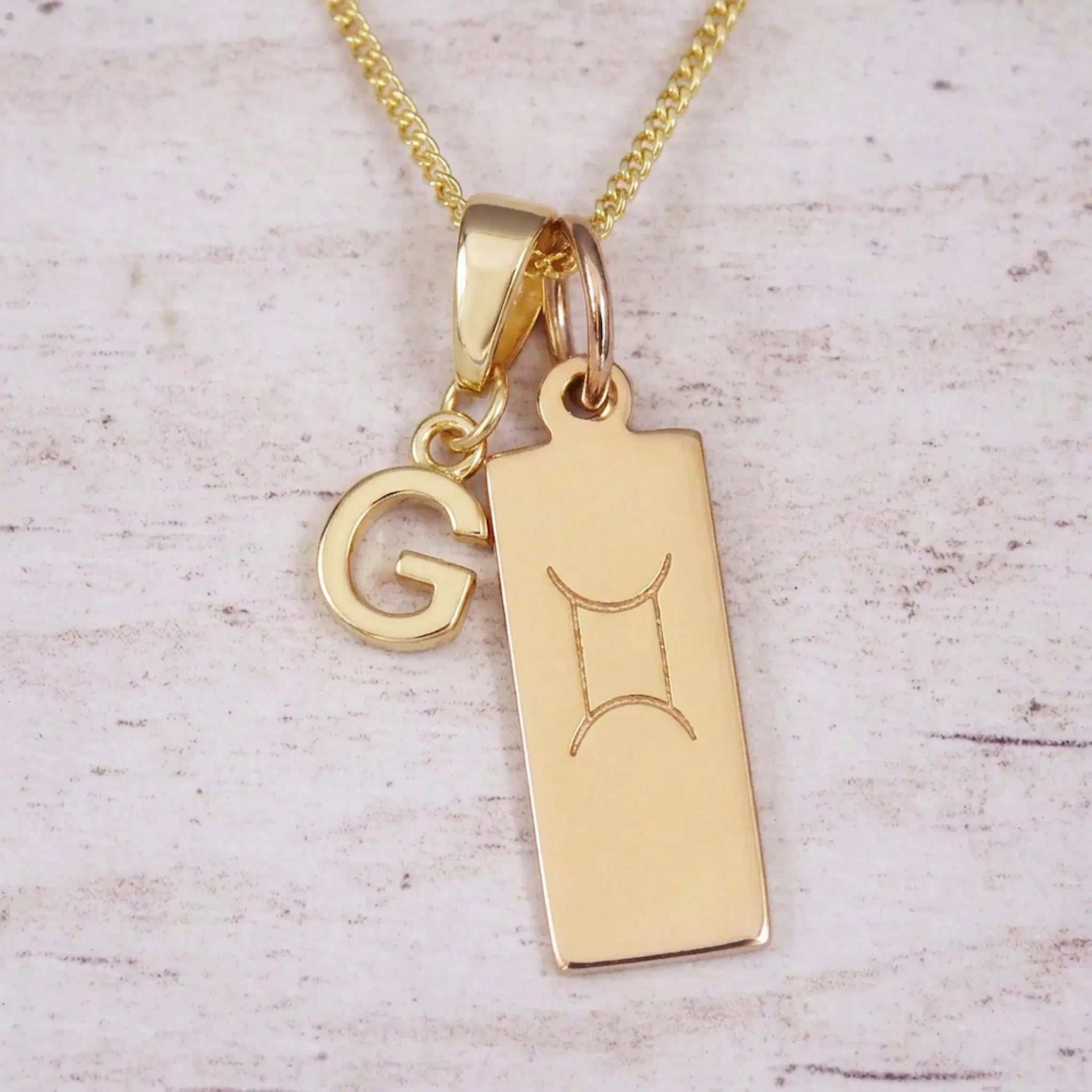 Gold initial G and Gemini zodiac bar necklace, close-up of the letter charm and engraved pendant on whitewashed wood